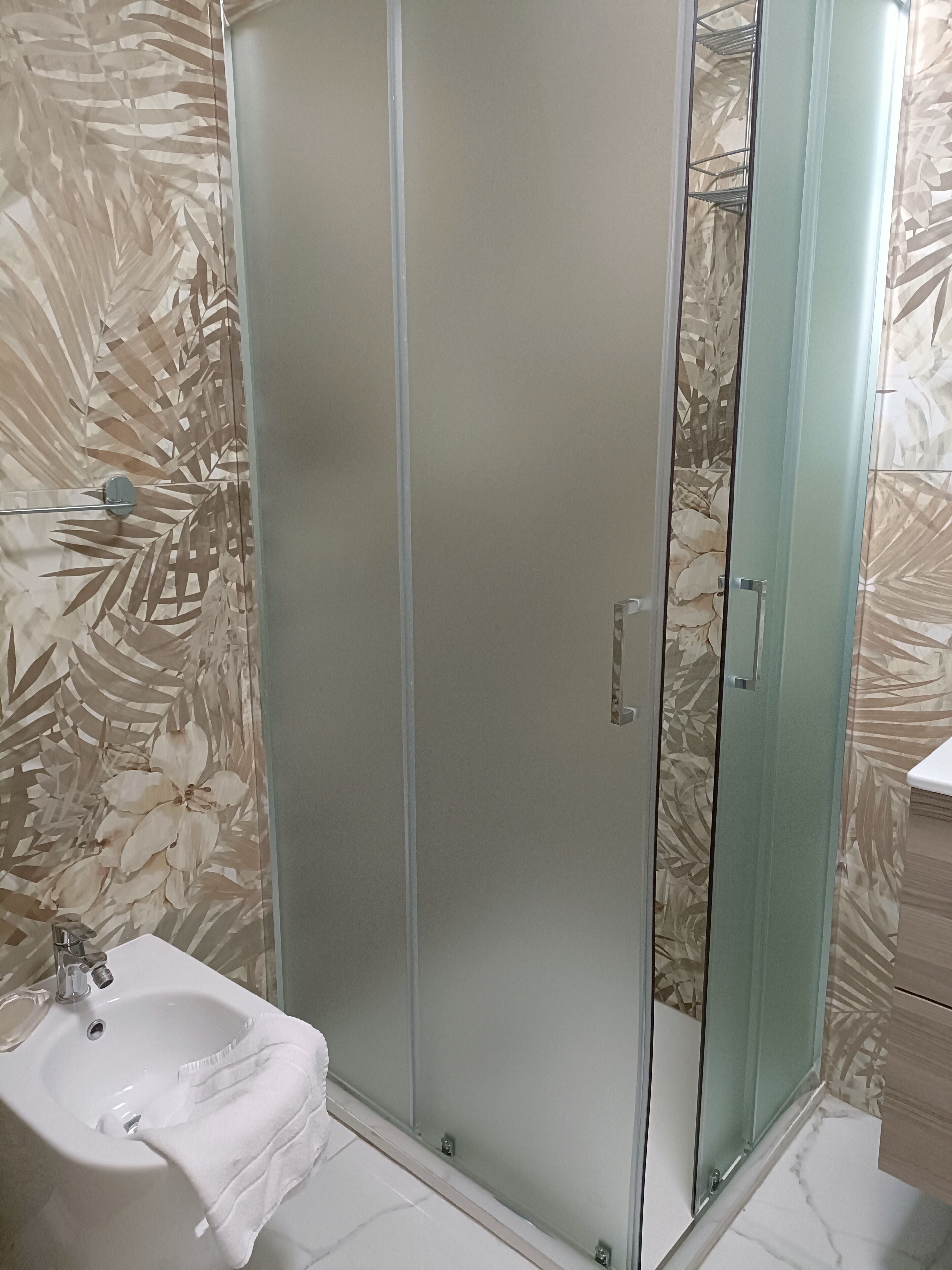 A bathroom with a glass shower enclosure, a sink with a towel, and patterned wallpaper.