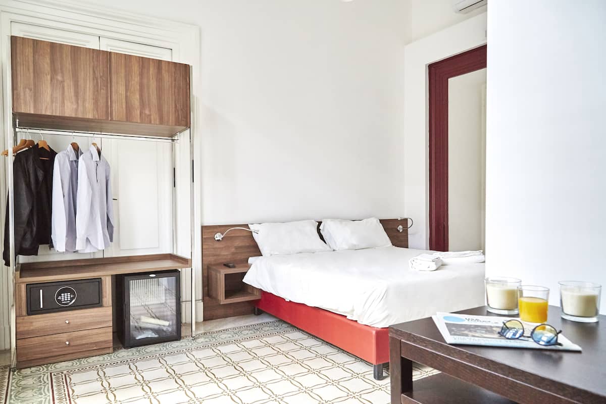 Habitación de hotel con cama, armario, mesita de noche con vasos y periódico, y un mueble pequeño.