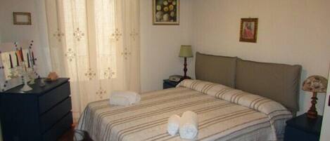 A bedroom with a bed, a nightstand, a dresser, a lamp, and a window with curtains.