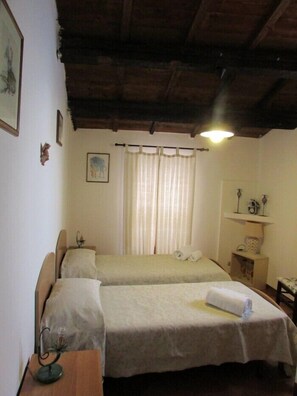 A room with two beds, a wooden ceiling, a window with curtains, and a small table with a lamp. - Il Balcone di Gilda (Cefalù)