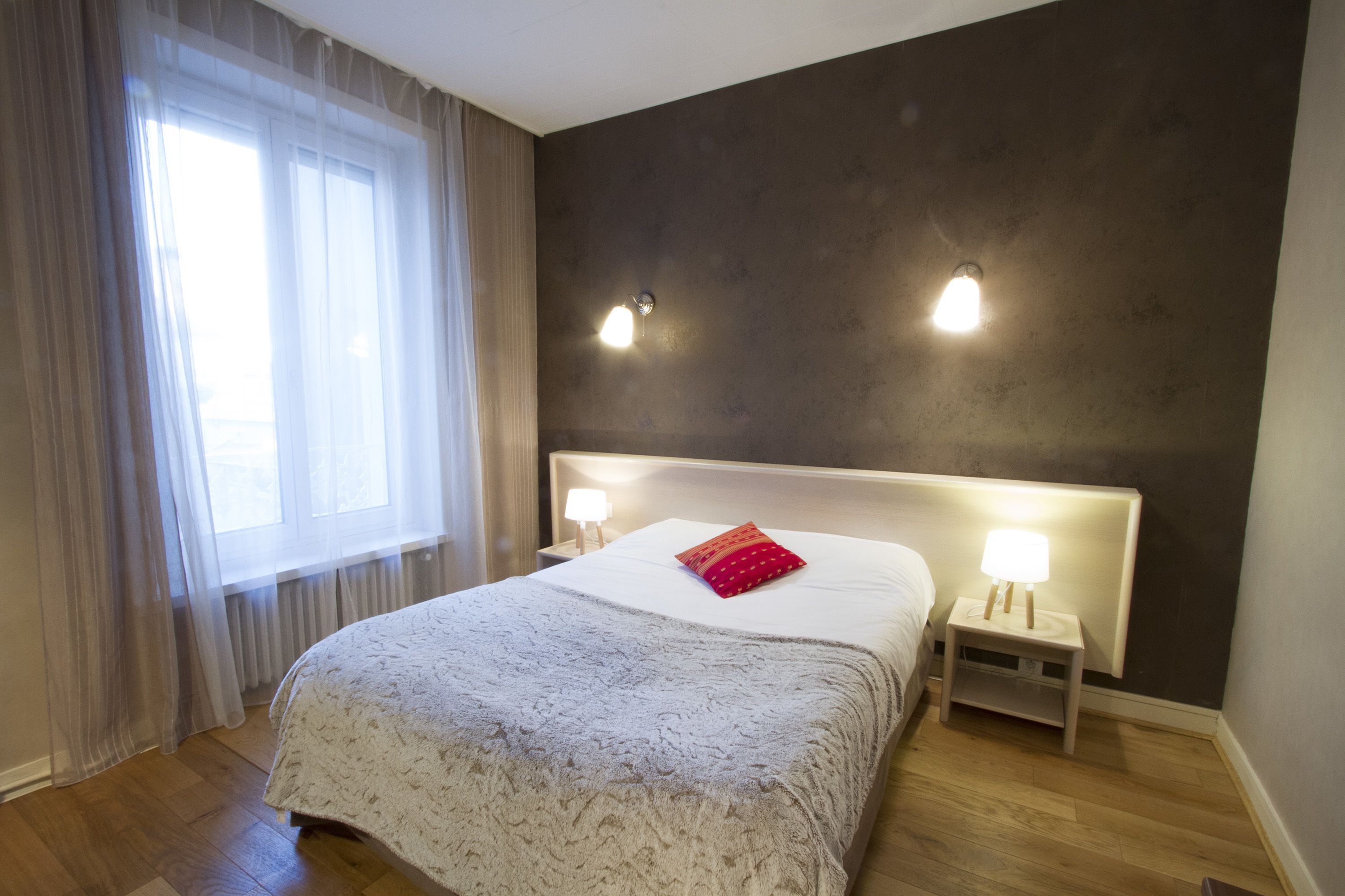 Una cama bien tendida con una colcha estampada y un almohárraga roja, flanqueada por dos mesitas de noche con lámparas, en una habitación con un ventanal grande y una pared oscura.