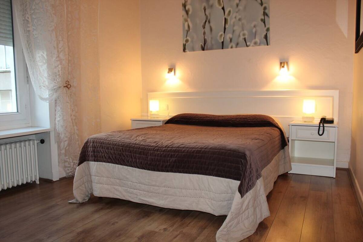 Une chambre à coucher avec un grand lit, deux tables de chevet avec des lampes, un radiateur et une fenêtre avec des rideaux.