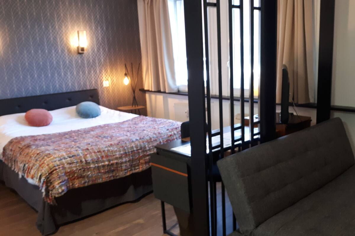 Une chambre à coucher avec un lit, un canapé, une fenêtre avec des rideaux et un luminaire fixé au mur.