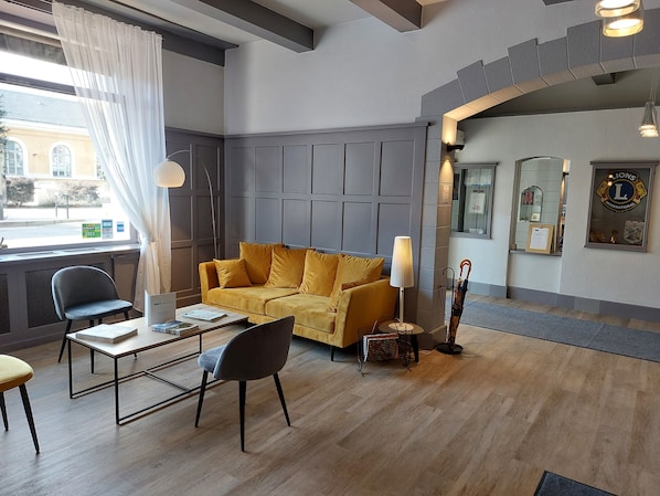 Un hall de hotel con un sofá amarillo, una mesa con sillas, una lámpara y una recepción.