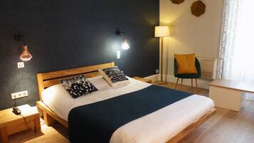 Ein ordentlich bezogenes Bett mit dunkelblauer Bettdecke und zwei Kissen, ein Nachttisch mit Lampe sowie ein Sessel in einem modernen Zimmer mit Holzboden und dunkler Wand.