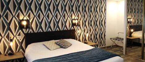 Una habitación de hotel moderna con una pared de estampado geométrico, una cama grande con cabecera oscura, dos mesitas de noche con lámparas y una mesita auxiliar con lámpara.