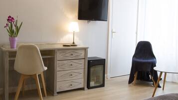 A room with a wooden desk, a chair, a television mounted on the wall, a small refrigerator, and a potted plant.