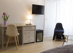 Una habitación con un escritorio de madera, una silla, un televisor en la pared, un refrigerador pequeño y una planta en maceta.