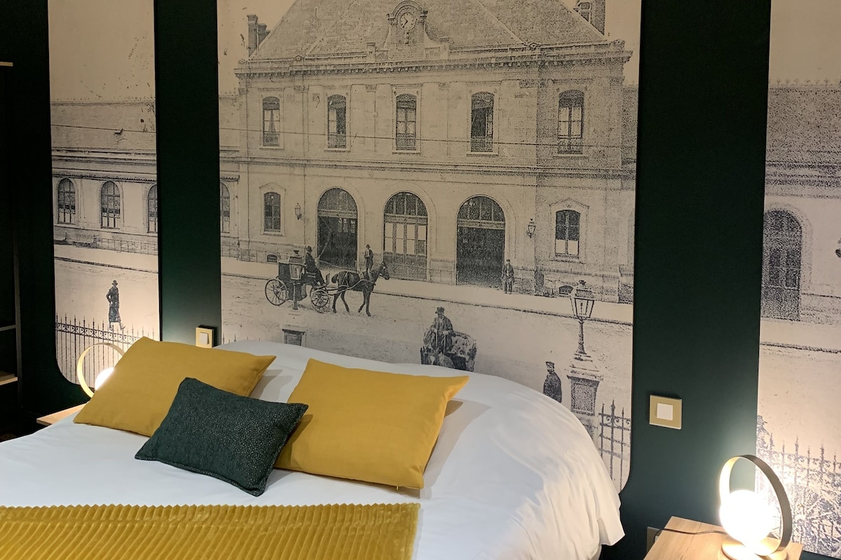 Una cama con sábanas blancas y almohadas mostaza, una mesita de noche con lámpara, y un mural en la pared de un edificio con gente y caballos.