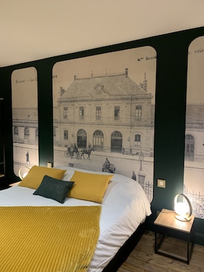 Una cama con sábanas blancas y almohadas mostaza, una mesita de noche con lámpara, y un mural en la pared de un edificio con gente y caballos.