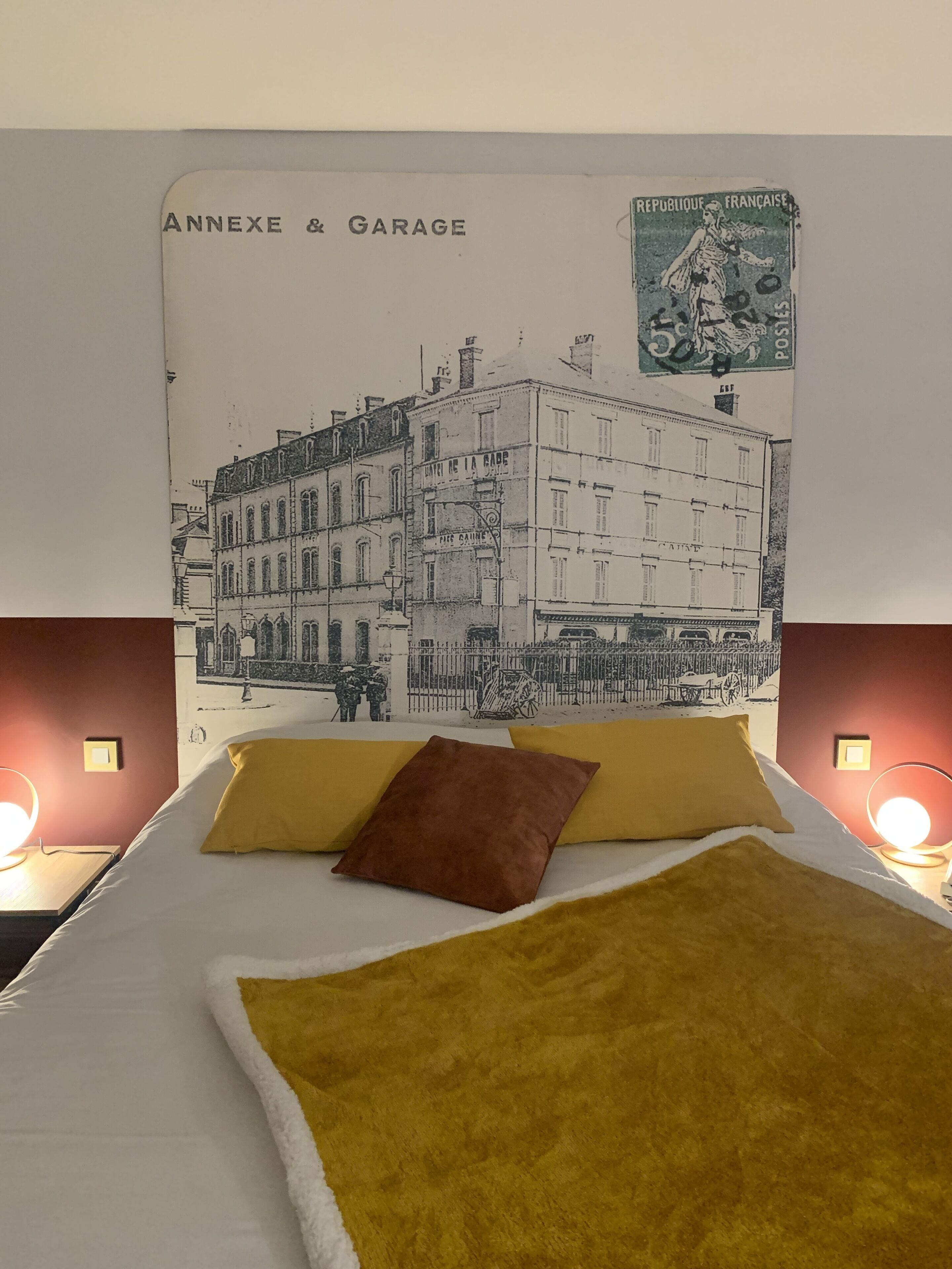 Un lit avec un oreiller jaune et marron, une couverture blanche et une affiche accrochée au mur représentant un bâtiment avec l’inscription « ANNEXE ET GARAGE ».