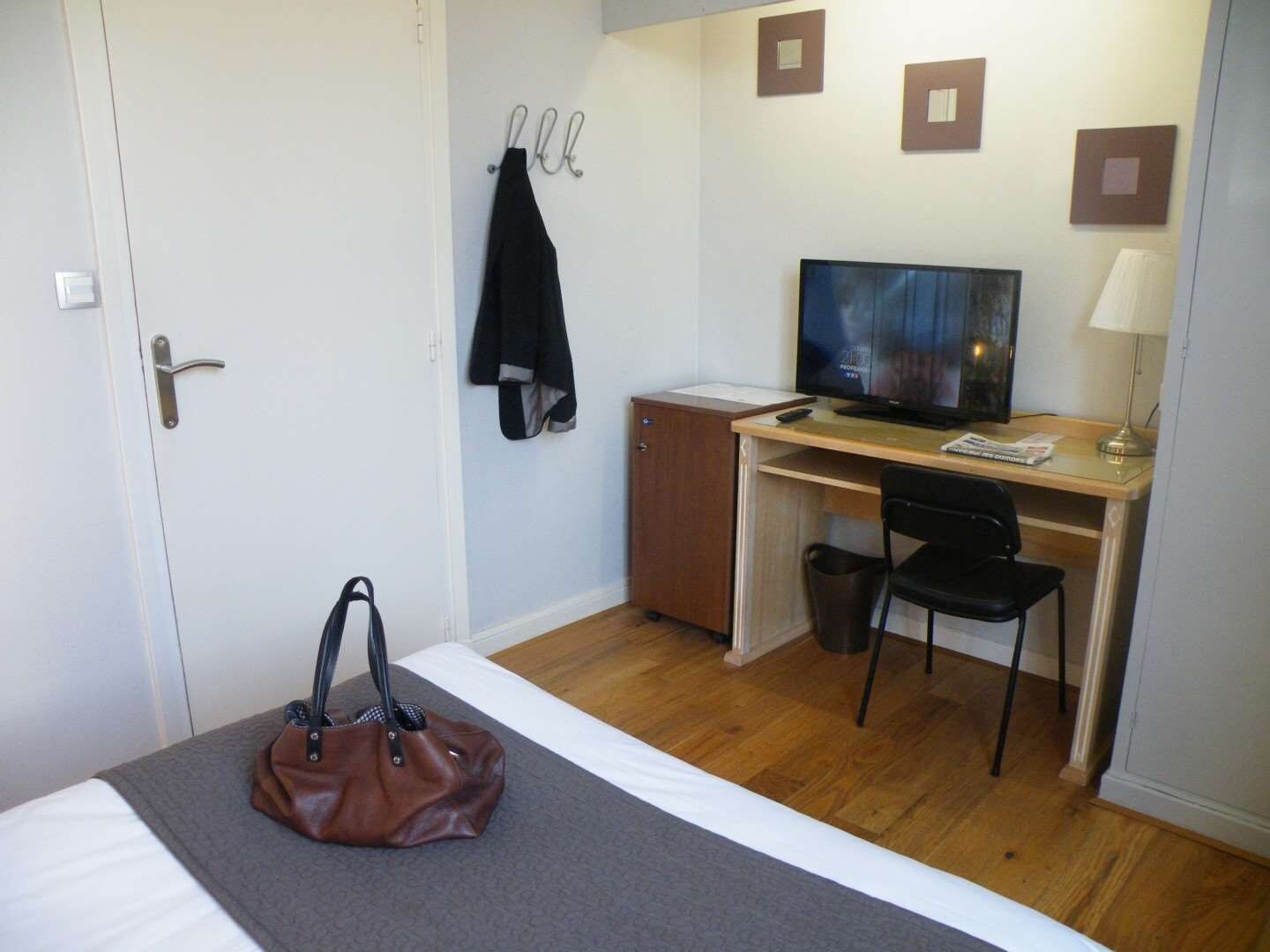 A hotel room with a bed, a desk with a television, a chair, a desk lamp, and a handbag on the bed.