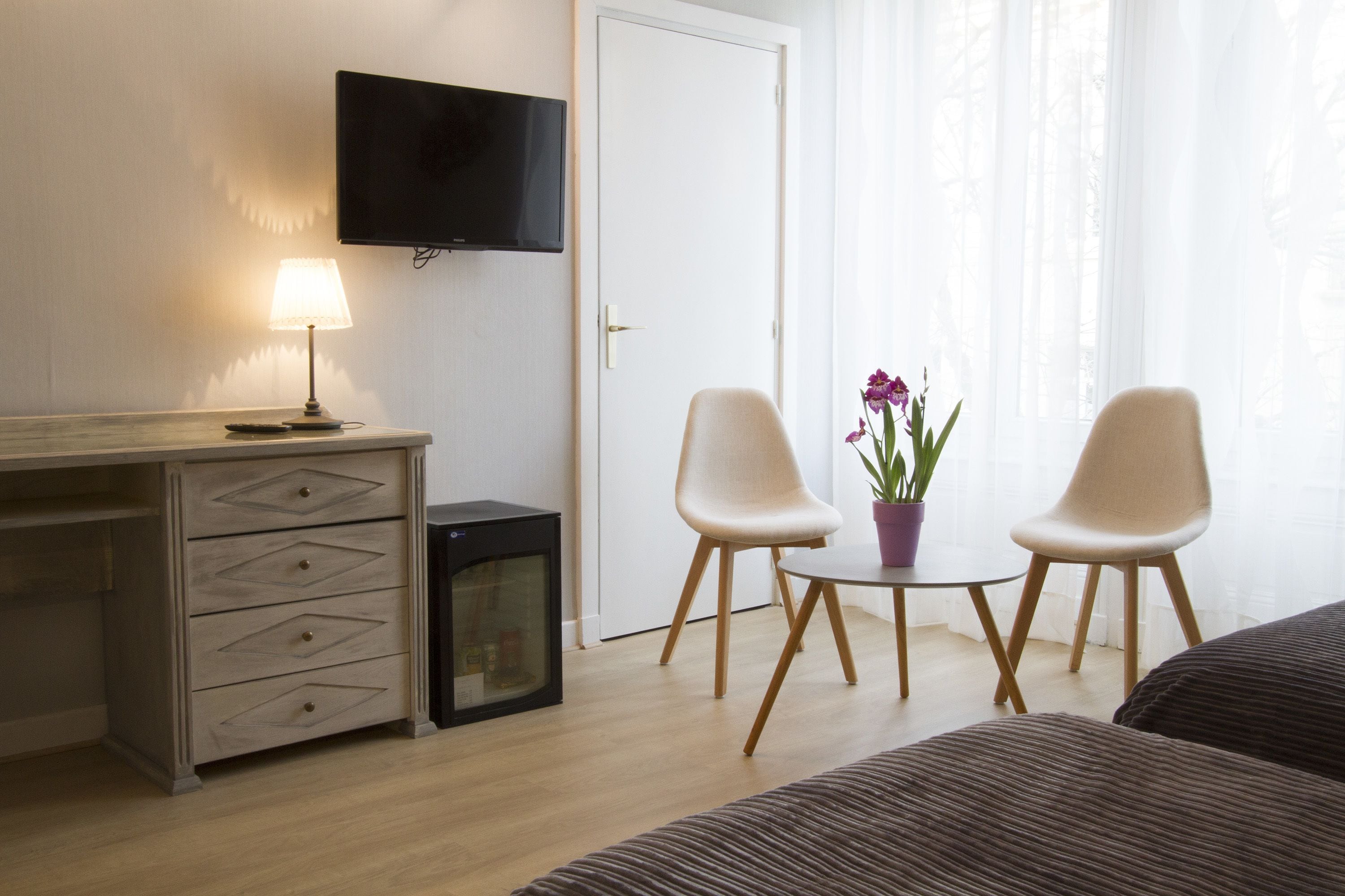Habitación de hotel con un escritorio de madera, un televisor montado en la pared, dos sillas, una mesita con una planta en maceta y una cama con ropa de cama a rayas.
