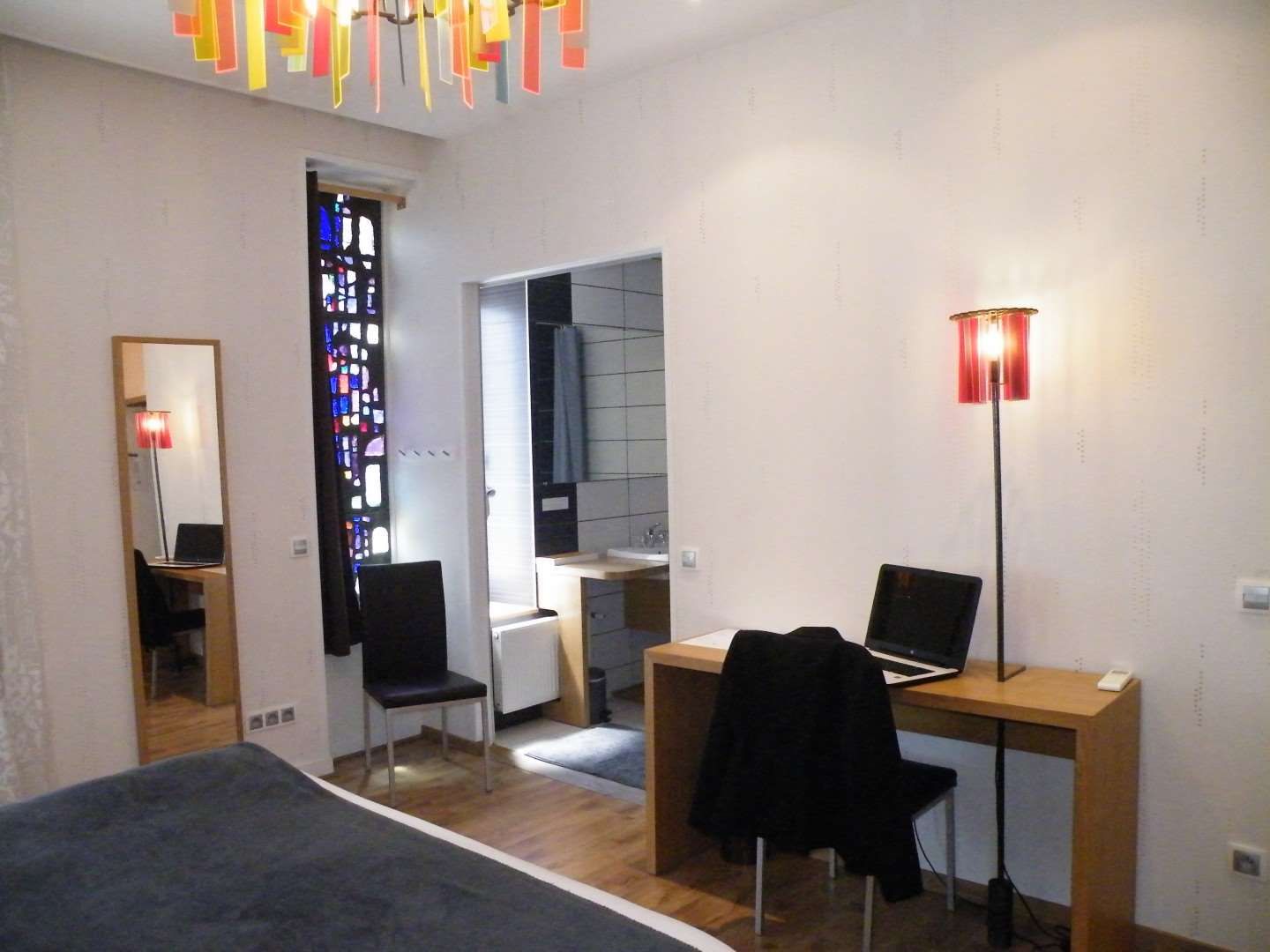 Un dormitorio con una cama, un escritorio con dos computadoras portátiles, una silla, un espejo y una ventana con un diseño de vitral.