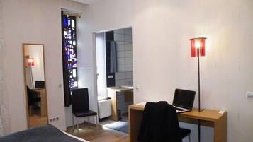 Un dormitorio con una cama, un escritorio con dos computadoras portátiles, una silla, un espejo y una ventana con un diseño de vitral.