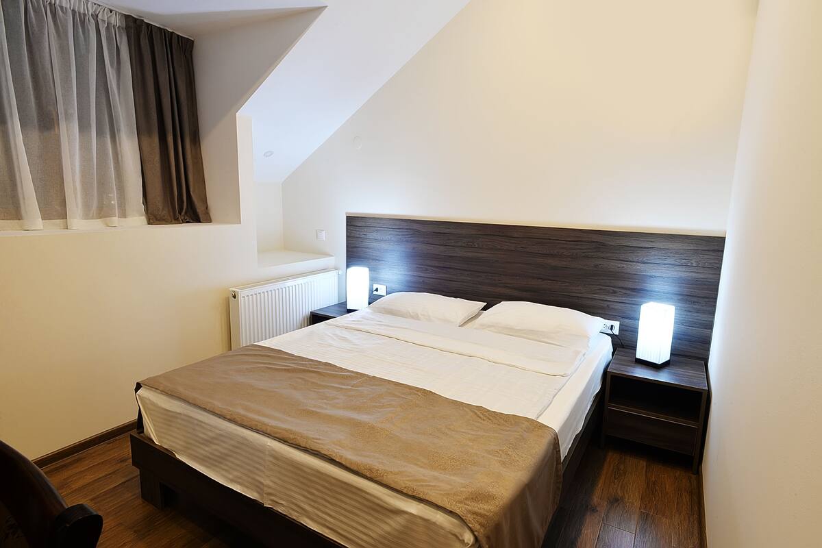 A neatly made bed with a wooden headboard, a bedside table with a lamp, and a window with curtains.