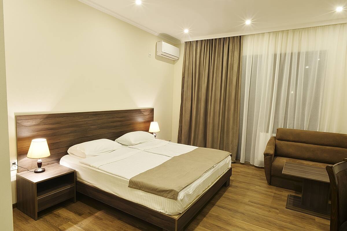 A hotel room with a wooden headboard bed, two bedside lamps, a brown armchair, and a large window with curtains.