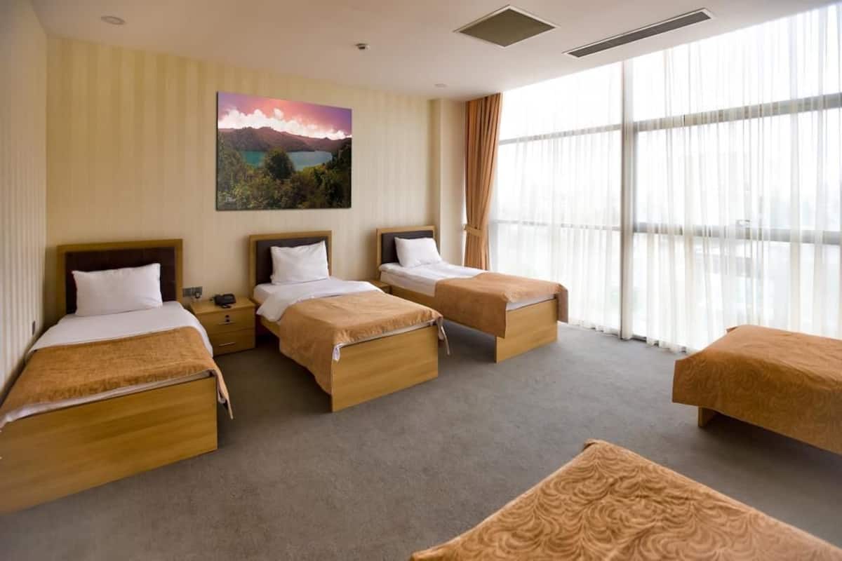 Une chambre d’hôtel avec quatre lits simples, une grande fenêtre avec des rideaux légers et un tableau représentant un paysage accroché au mur.