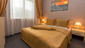 A hotel room with a large bed, two bedside tables with lamps, a cityscape painting on the wall, and sheer curtains. - Riva Hotel (Baku)