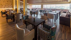 A spacious indoor area with multiple tables and chairs, large windows offering a city view, and a wooden ceiling. - Riva Hotel (Baku)