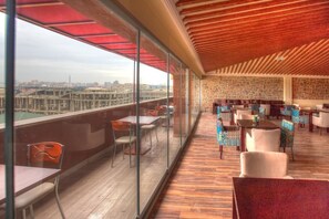 Un vaste balcon offrant une vue sur le paysage urbain, aménagé avec des tables et des chaises, et un mur en pierre orné d’un motif géométrique.