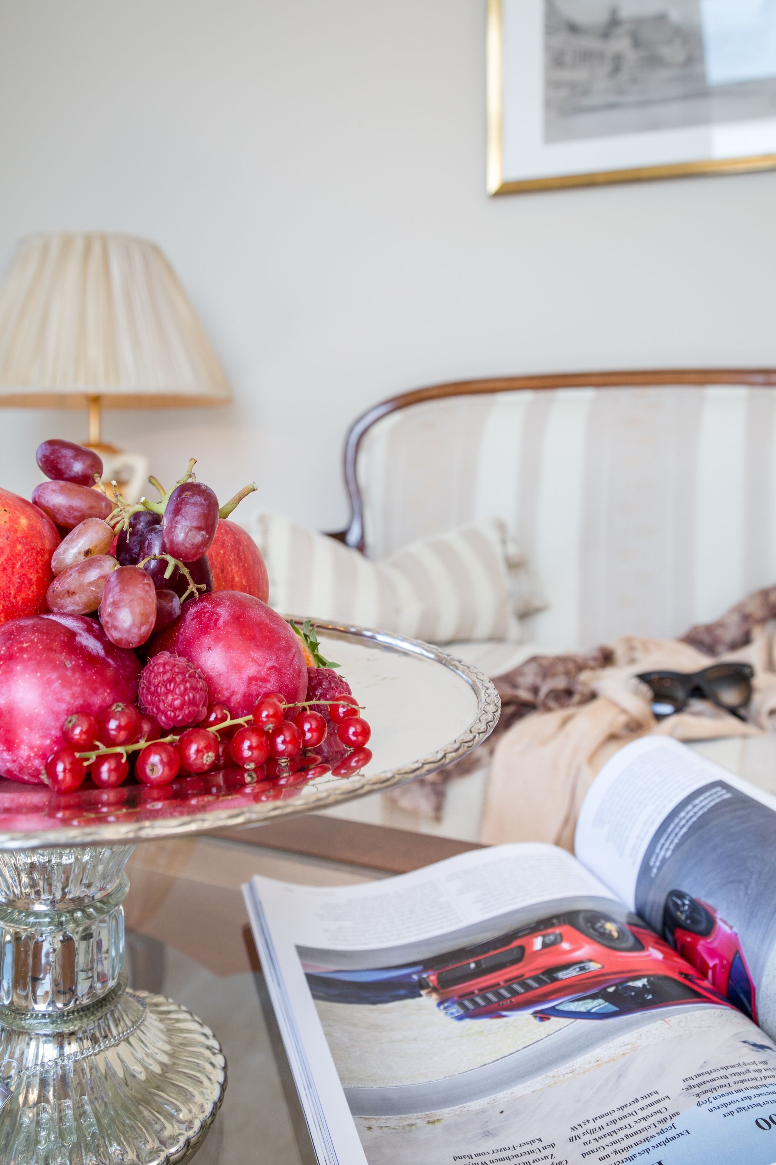 Une corbeille de fruits posée sur un présentoir en verre, un lit avec une literie à rayures, une lampe et un magazine illustré d’une photo de voiture.