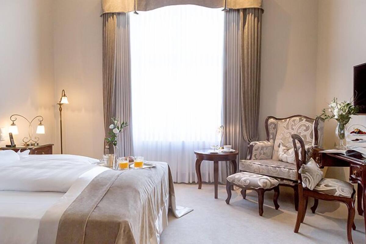 A hotel room with a large bed, a desk with a chair, a small table with a lamp, and a window with curtains.