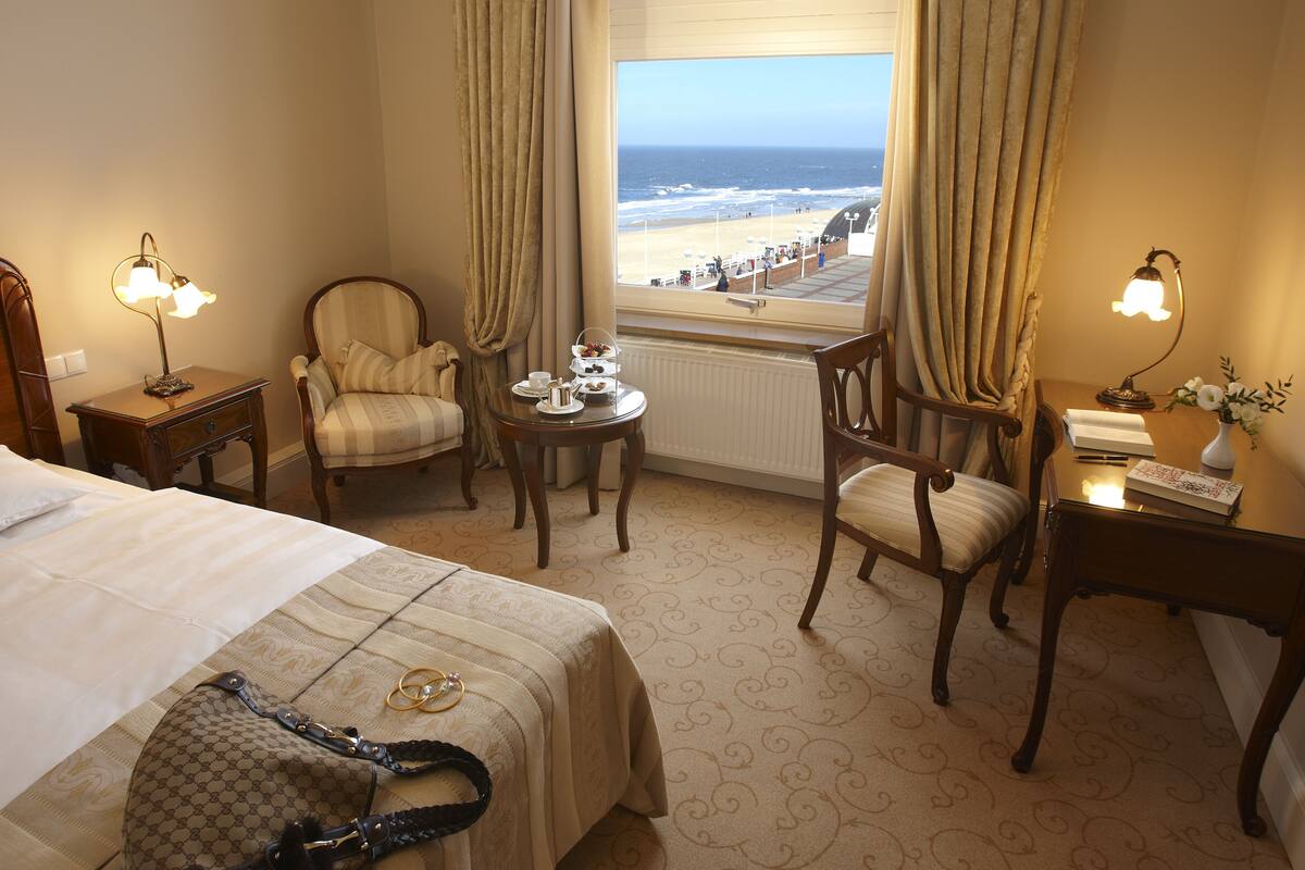 A hotel room with a bed, two armchairs, a small table with a tray of tea, a desk with a lamp, and a view of the beach.