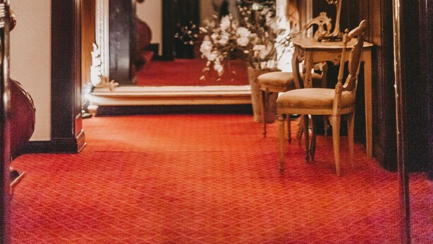 Un amplio pasillo con alfombra roja, candelabros ornamentales y un espejo que refleja una escalera.