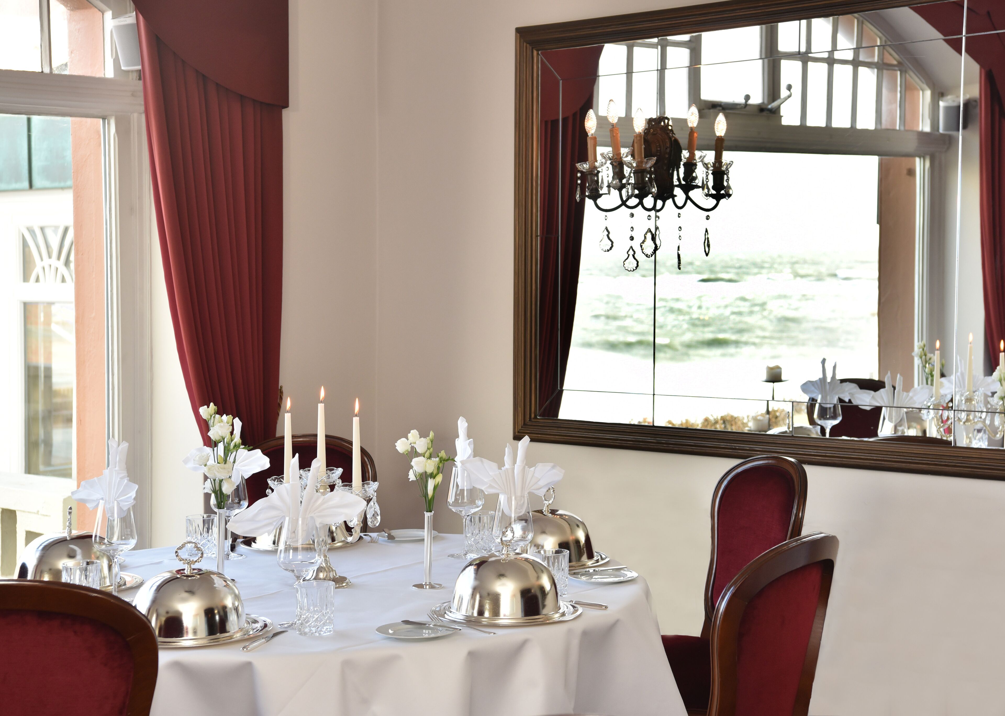 A dining table set with white tablecloth, silverware, and candles, with a view of the sea through a window.
