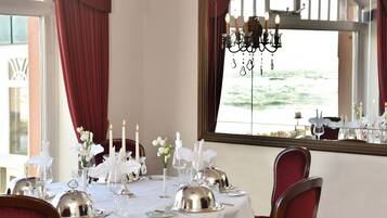 A dining table set with white tablecloth, silverware, and candles, with a view of the sea through a window.