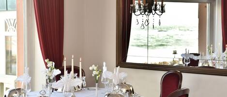 Une table à manger dressée avec une nappe blanche, des couverts en argent et des bougies, offrant une vue sur la mer par la fenêtre.