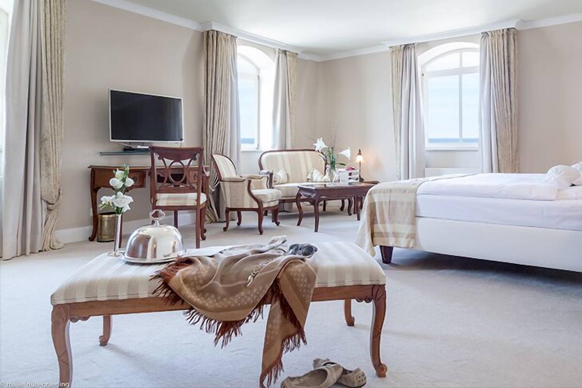 A hotel room with a bed, a desk with a television, a chair, a bench, and a window with curtains.