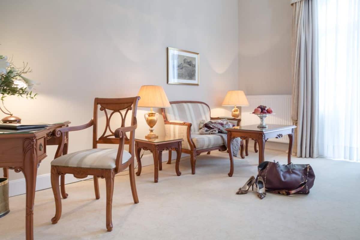 A hotel room with a wooden chair, a sofa, a small table with a lamp, a radiator, and a window with curtains.