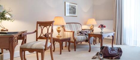 A hotel room with a wooden chair, a sofa, a small table with a lamp, a radiator, and a window with curtains.