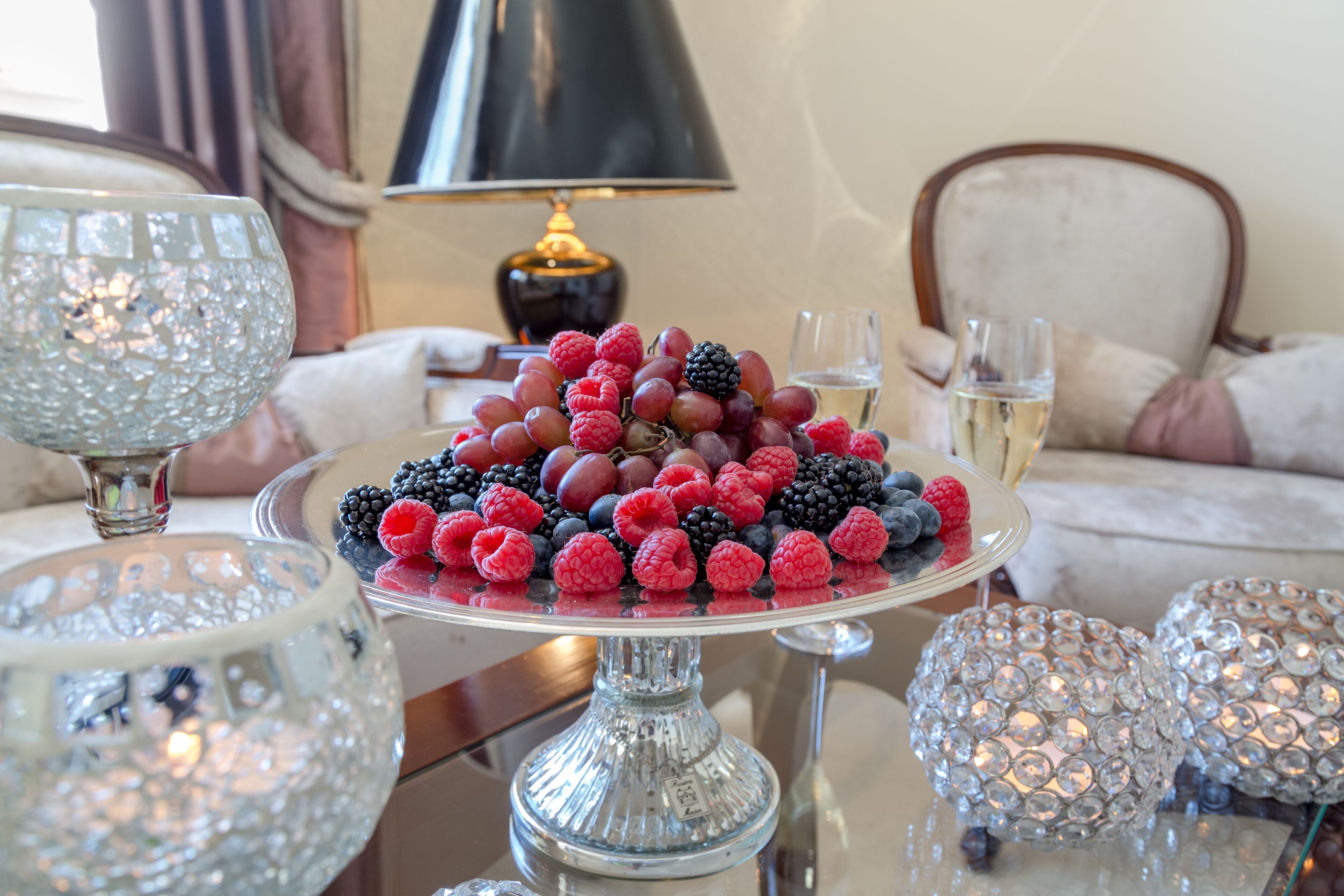 A dining table with a fruit platter, crystal glasses, and a lamp in the background.