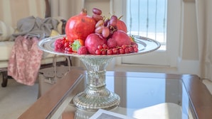 Una mesa de comedor con un plato de fruta, una revista y vista al mar a través de una puerta de balcón.
