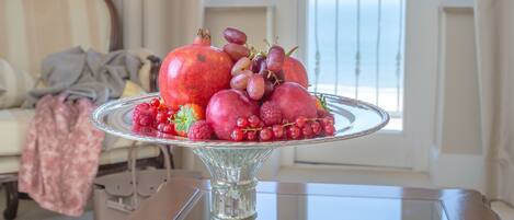 A dining table with a fruit platter, a magazine, and a view of the sea through a balcony door.