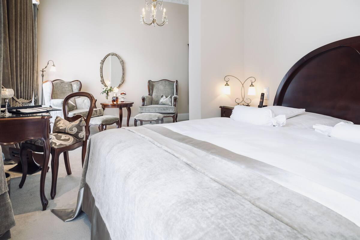 A hotel room with a large bed, two armchairs, a desk, a mirror, and a chandelier.