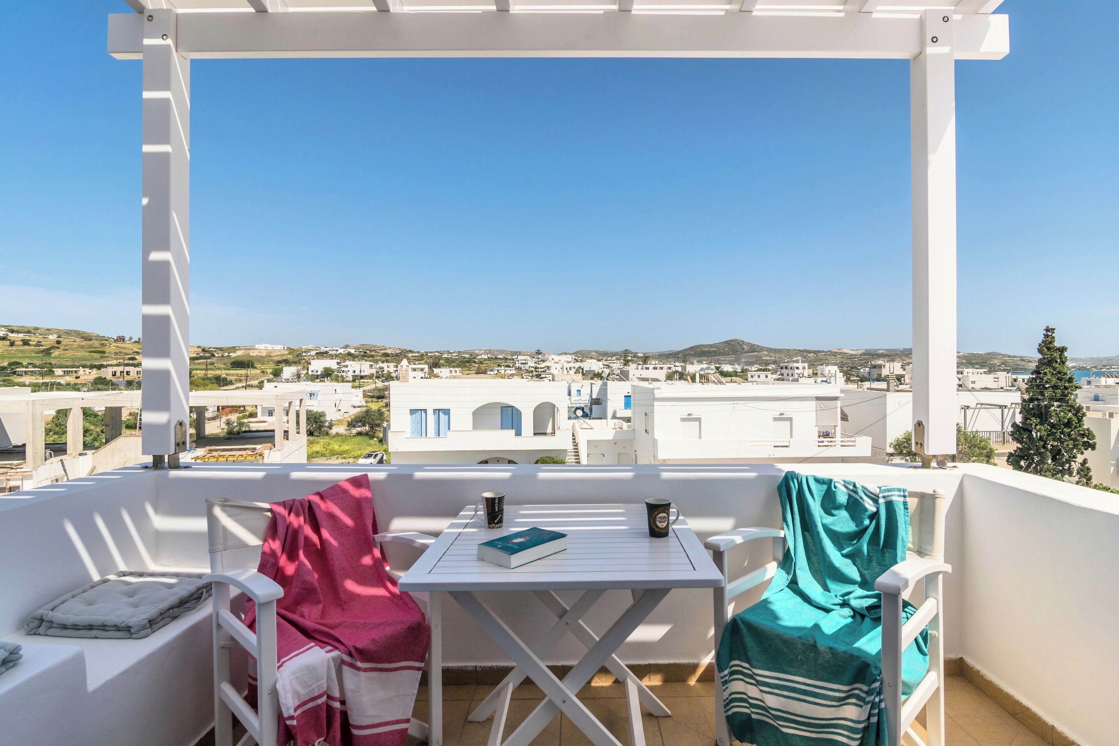A balcony with a white table and chairs, a book, and two cups. The view includes white buildings and a clear blue sky.
