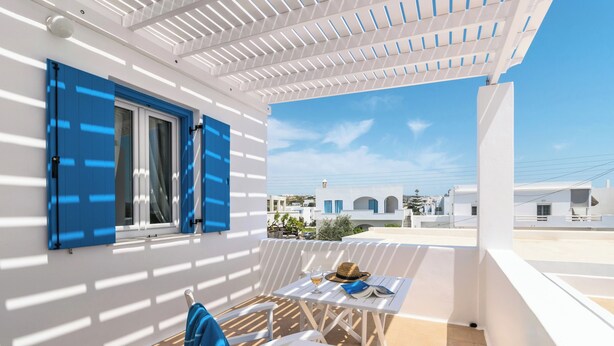 A balcony with a white pergola, a table with a hat and a glass, and a chair. - Livanios Studios (Milos)