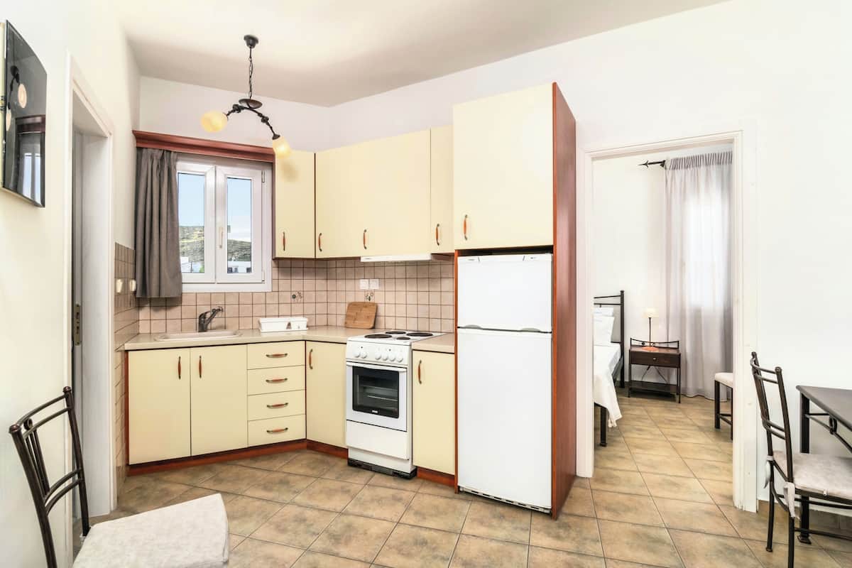 A compact kitchen with beige cabinets, a white refrigerator, a stove, and a sink. There is a dining table with chairs and a window with curtains.