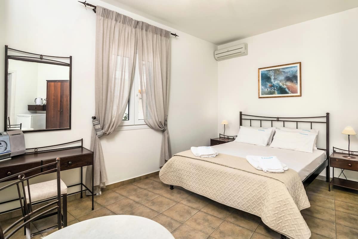 A hotel room with a bed, a desk, a chair, a mirror, a window with curtains, an air conditioner, and a painting on the wall.