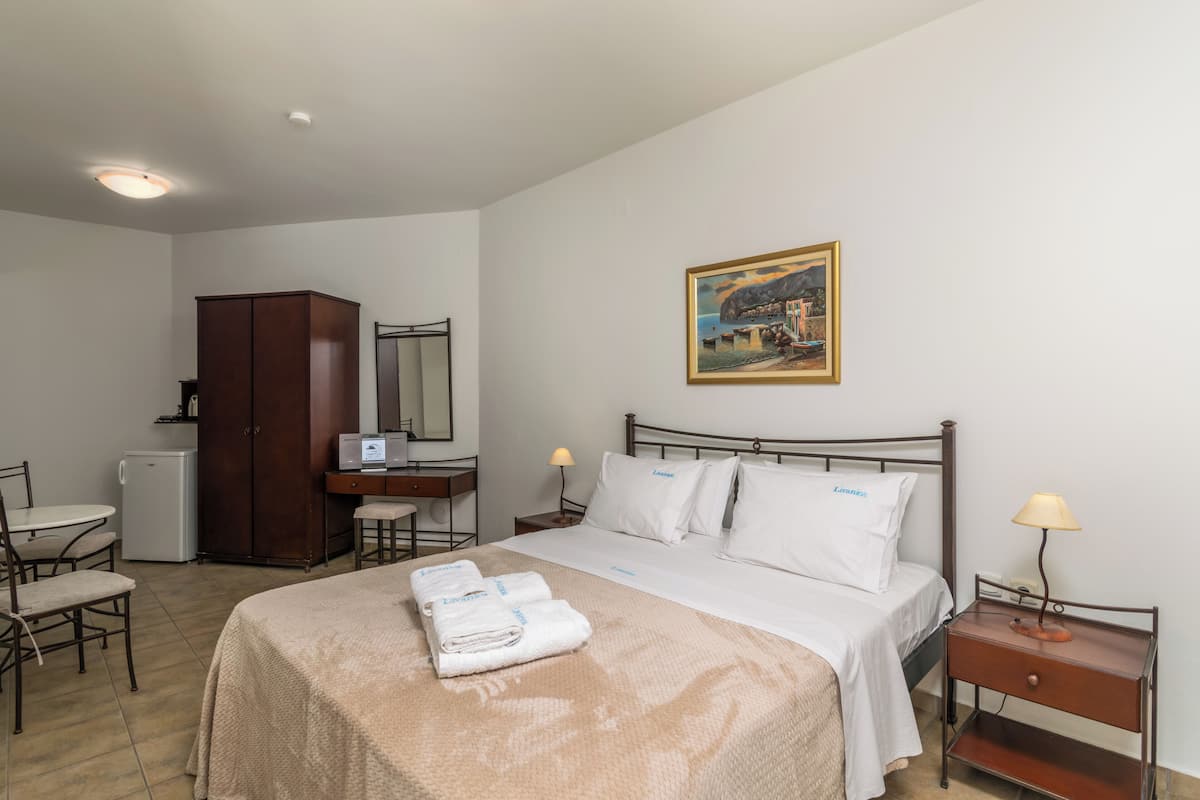 A hotel room with a bed, bedside table, lamp, desk, chair, wardrobe, and a painting on the wall.