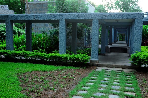 A covered walkway with stone columns, surrounded by lush greenery and a well-maintained garden.