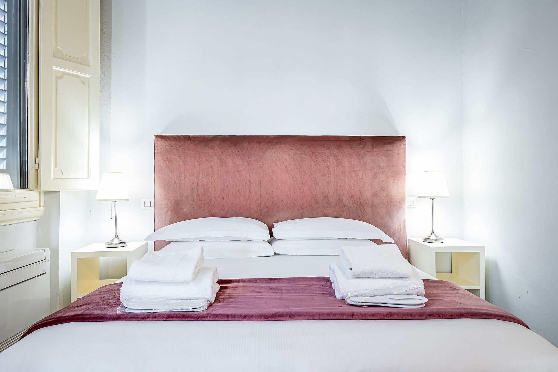 Ein ordentlich bezogenes Bett mit weißen Leinentüchern, einem pinkfarbenen Kopfteil und zwei Nachttischlampen.