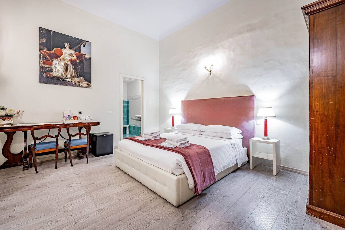 A hotel room with a bed, a desk, a chair, a wardrobe, and a painting on the wall.