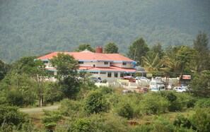 A multi-story building with a red roof, surrounded by lush greenery and parked vehicles.