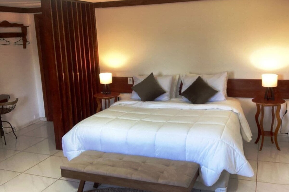 A hotel room with a large bed, two bedside tables, a bench at the foot of the bed, and a wall-mounted shelf.