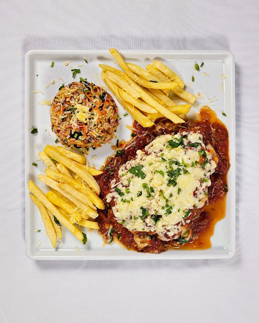 A square plate with fried rice, French fries, and a meat dish with cheese and herbs.
